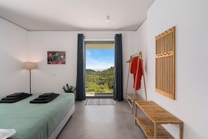 Ein Schlafzimmer mit Bett, Nachttisch, Pendelleuchte, einem Gemälde, einem Fenster mit Vorhängen und Blick ins Grüne.