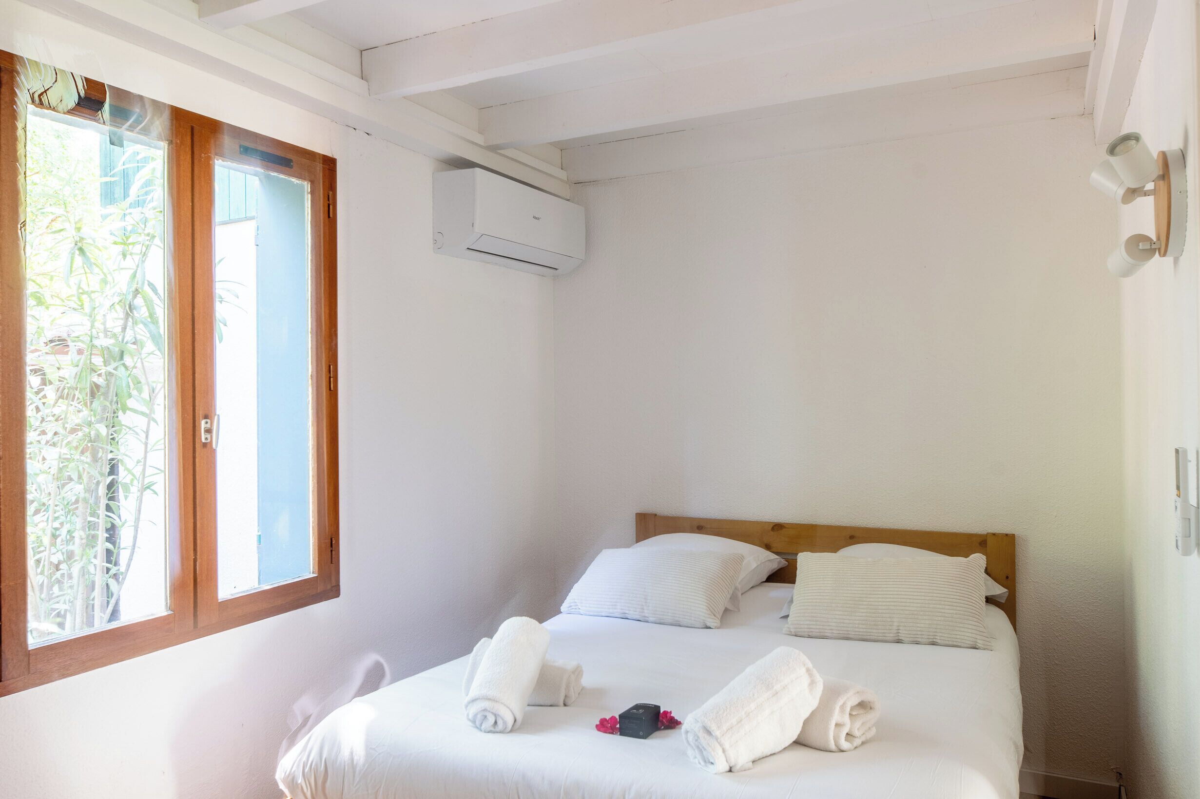 Habitación de hotel con cama, dos toallas enrolladas, ventana con vista a follaje y un equipo de aire acondicionado.