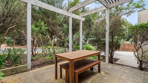 Un banc en bois, placé sous une pergola dans un jardin arboré, avec un chemin dallé.