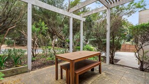 A wooden bench under a pergola in a garden with trees and a paved path. - 9 Doors From Front Beach (POINT LONSDALE)