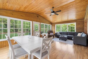 Ein geräumiger Raum mit Holzbalkendecke, großen Fenstern, einem Esstisch mit Stühlen, einem grauen Sofa und einem Deckenventilator.