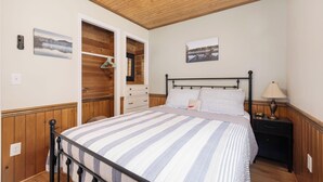 Een slaapkamer met een bed, houten plafond, nachtkastje met lamp, een wandklok en een deur naar een andere kamer.