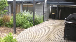A wooden deck with a barbecue grill, surrounded by plants and a fenced area. - Beachside Haven - Point Lonsdale (POINT LONSDALE)