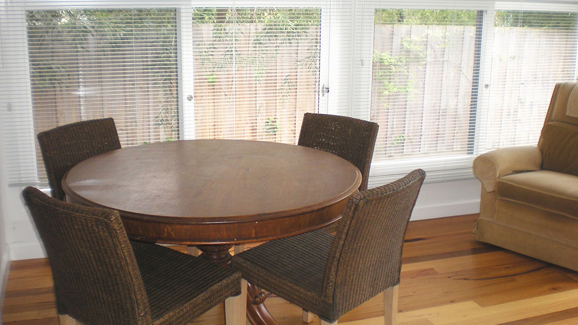 Een eethoek met een ronde tafel en vier stoelen, grote ramen met zonwering, en een bank.