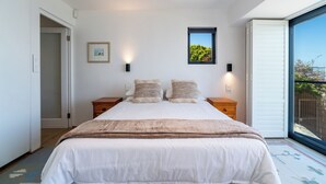 Un dormitorio con una cama grande, dos mesitas de noche, un ventanal con vistas y una puerta que da a otra habitación.
