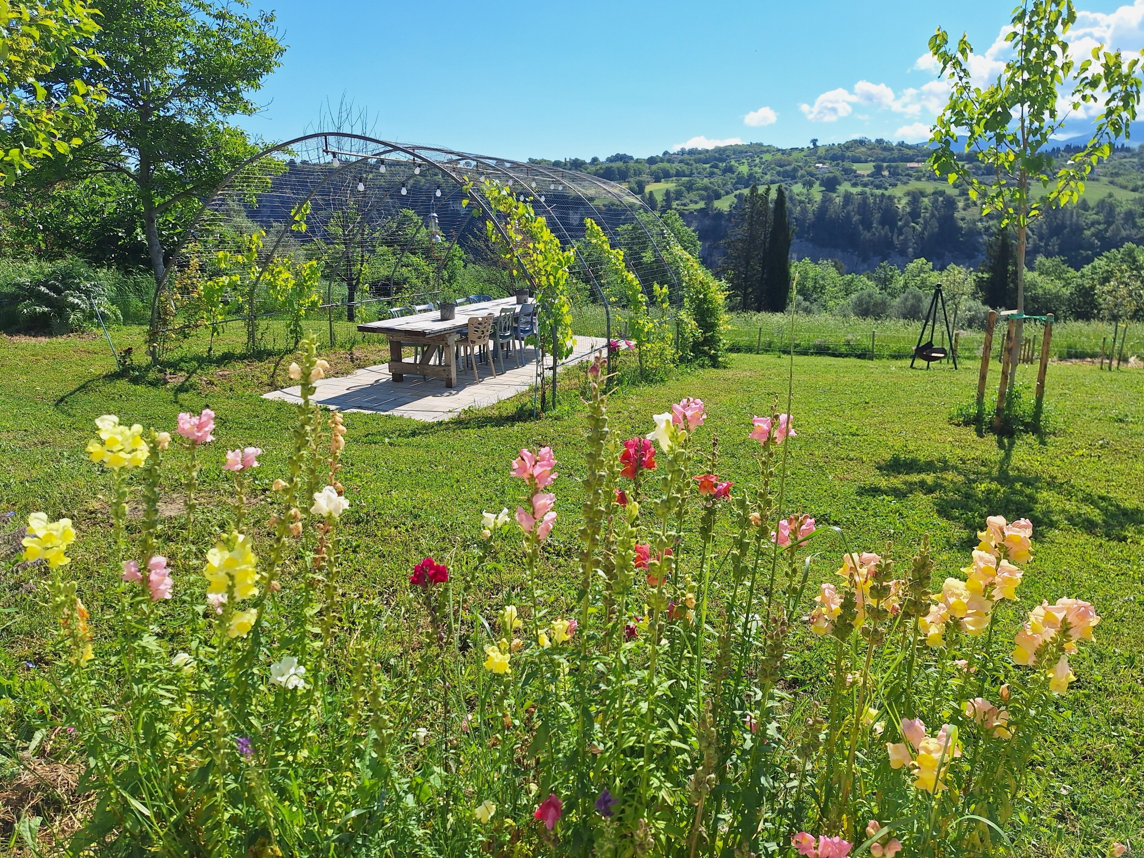 Un espace de restauration extérieur rustique, avec une table et des chaises, entouré d’un jardin luxuriant où poussent différentes plantes à fleurs.