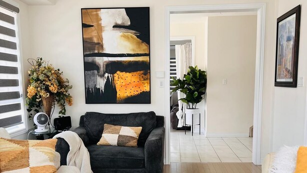 A modern living room with a black sofa, a white armchair, and a round coffee table with flowers. There is a large abstract painting on the wall and a framed picture above the coffee table. -     The Monarch Residence | Luxury for Families & Groups (Oshawa)