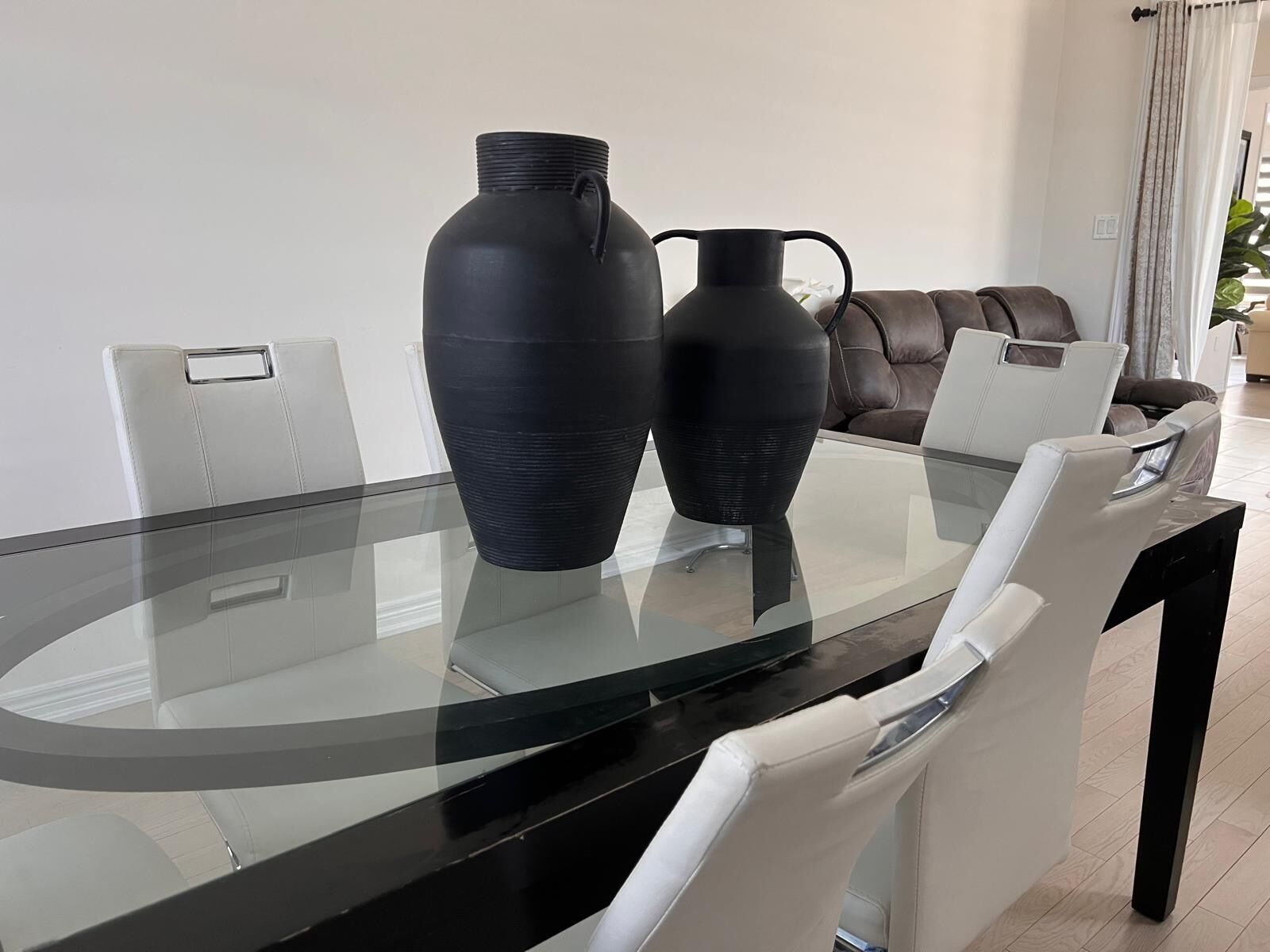A modern dining area with a glass table, two black vases, white chairs, and a brown leather sofa.