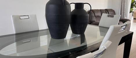 A modern dining area with a glass table, two black vases, white chairs, and a brown leather sofa.