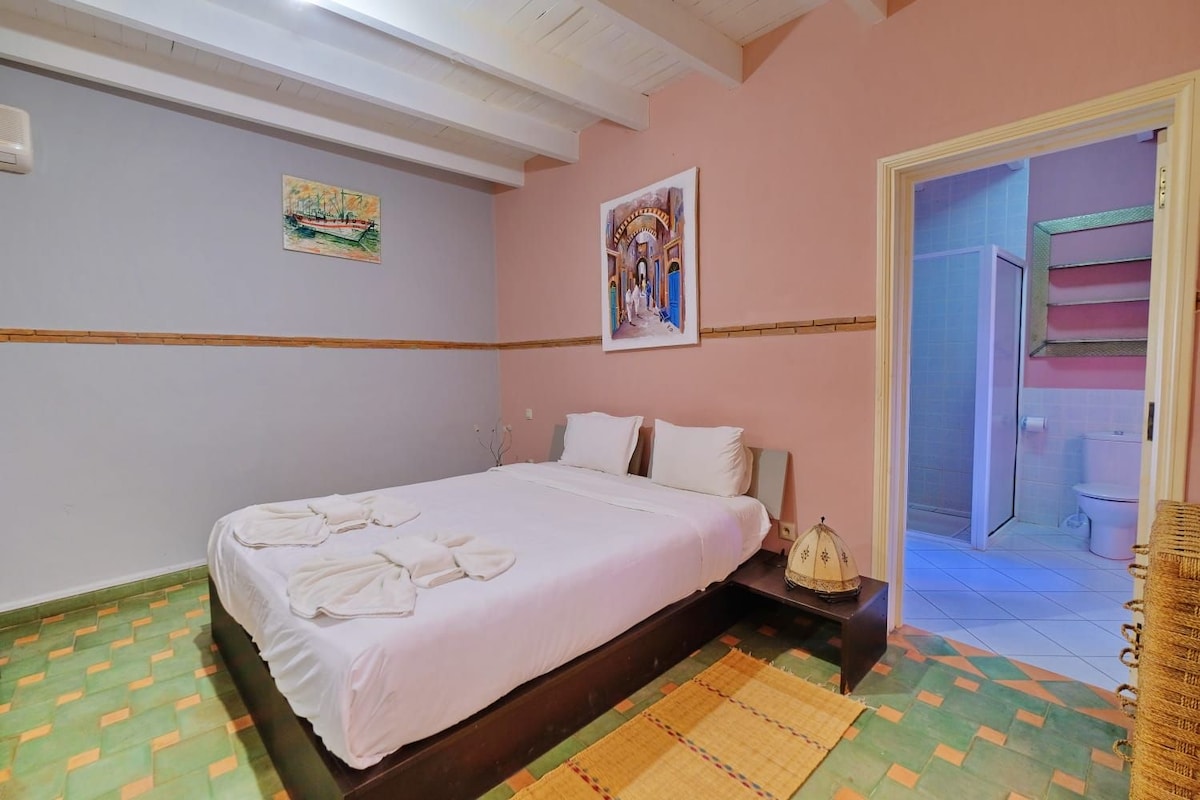 Une chambre à coucher comprenant un lit, une table de chevet, un climatiseur fixé au mur et une porte donnant sur une salle de bain.