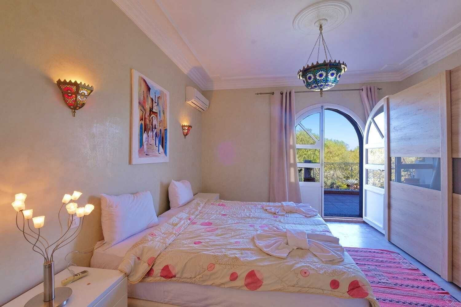 A bedroom with a bed, bedside lamps, a painting, an air conditioning unit, and a view through a window.