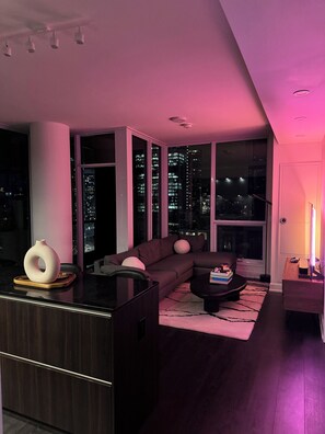Ein modernes Wohnzimmer mit dunklem Holzboden, einem großen Fenster mit Blick auf eine Skyline, einem dunkelbraunen Sofa und einem kleinen Couchtisch.