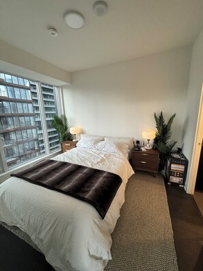 Een slaapkamer met een groot bed, twee nachtkastjes, een raam met uitzicht op gebouwen en een deur.