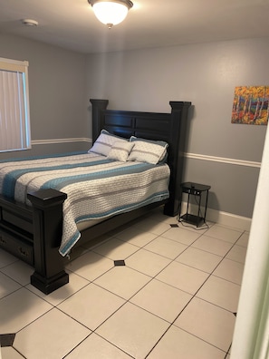 A bedroom with a bed, a nightstand, a window with blinds, and a wall-mounted painting.