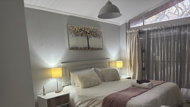 Un dormitorio con cama, dos lámparas de noche, mesita de noche, un cuadro y una ventana con cortinas.