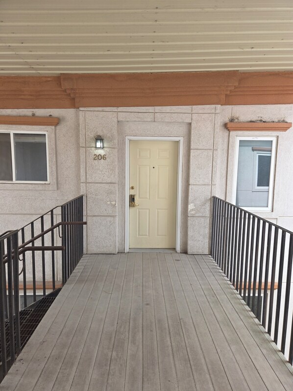 A covered wooden deck leading to a beige door with a number '206' above it, flanked by windows and a black metal railing.