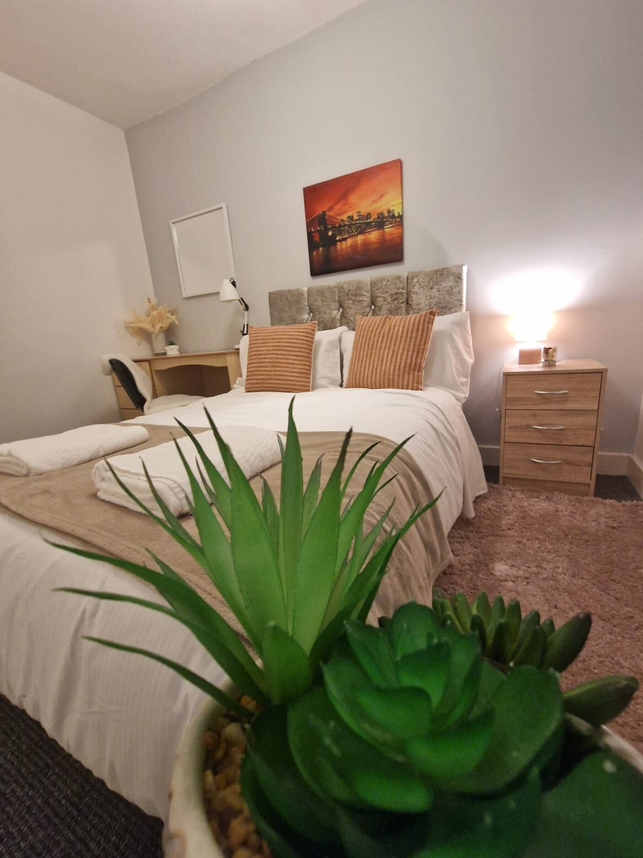 A bedroom with a bed, a nightstand, a lamp, a potted plant, and a painting on the wall.