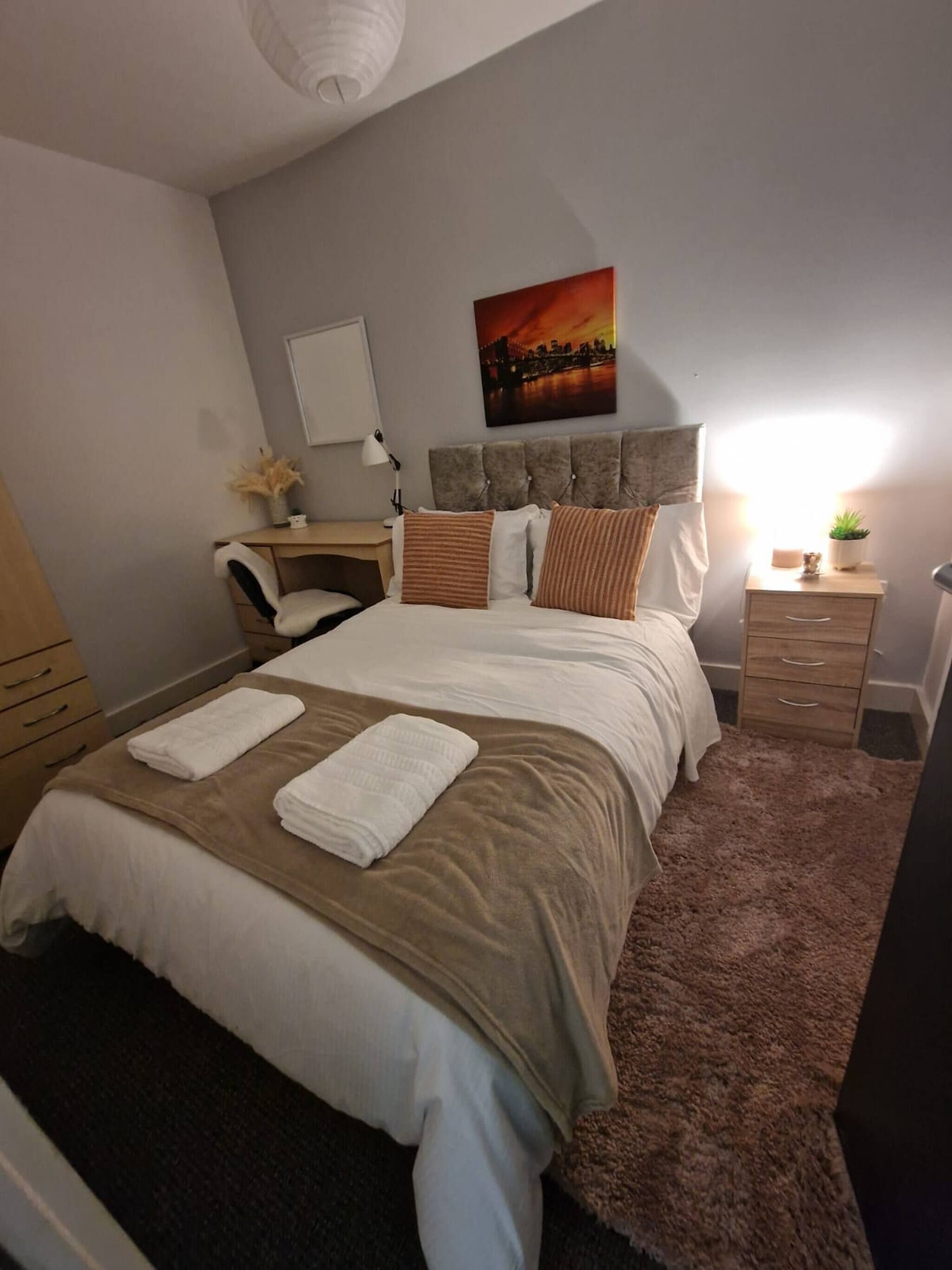 Un dormitorio ordenado con cama, mesitas de noche, una silla, un escritorio y un cuadro en la pared.