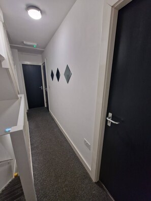 A hallway with a carpeted floor, a white wall with three diamond-shaped decorations, a black door, and a white door with a handle. - Luxury Home in Kensington for 21 Guests (Liverpool)
