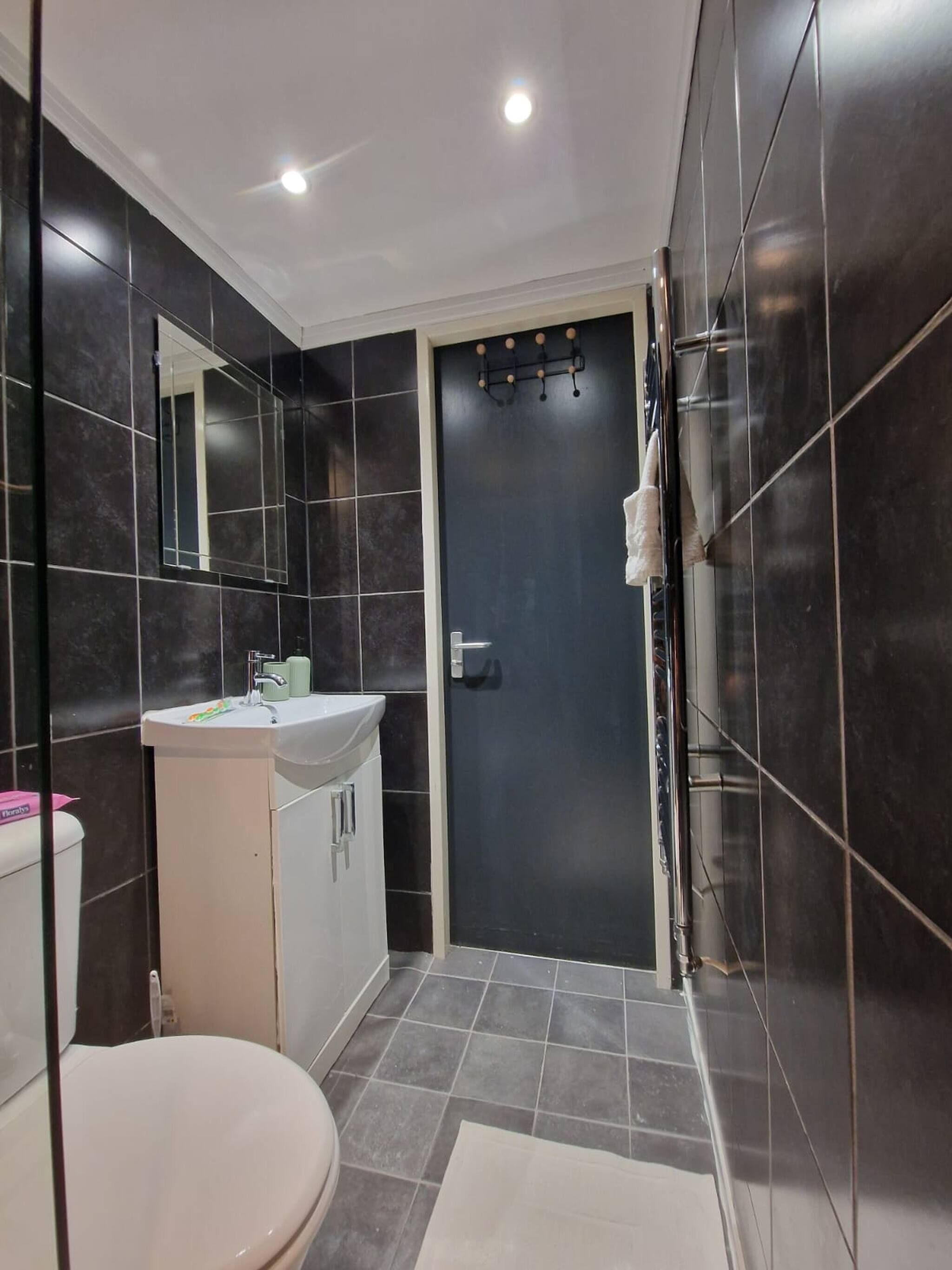 A bathroom with a toilet, sink, mirror, and dark tiled walls.
