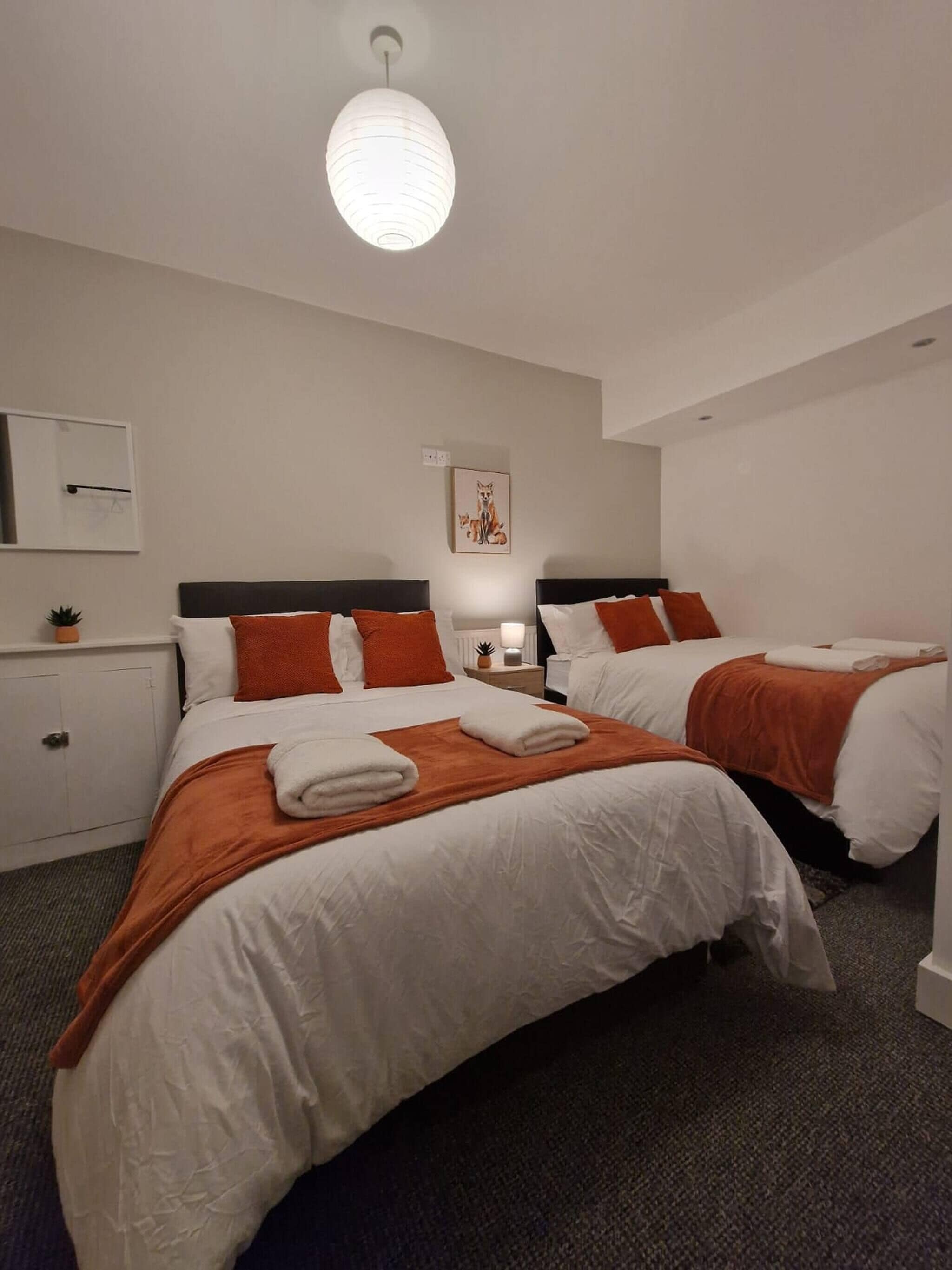 Habitación de hotel con dos camas, ropa de cama blanca, almohadones naranja, una lámpara blanca, un cuadro en la pared y una lámpara de techo.