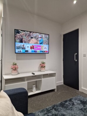 Una sala moderna con televisor de pantalla plana montado en la pared, un mueble blanco para entretenimiento con dos jarrones de flores, un sofá azul oscuro y una puerta negra.