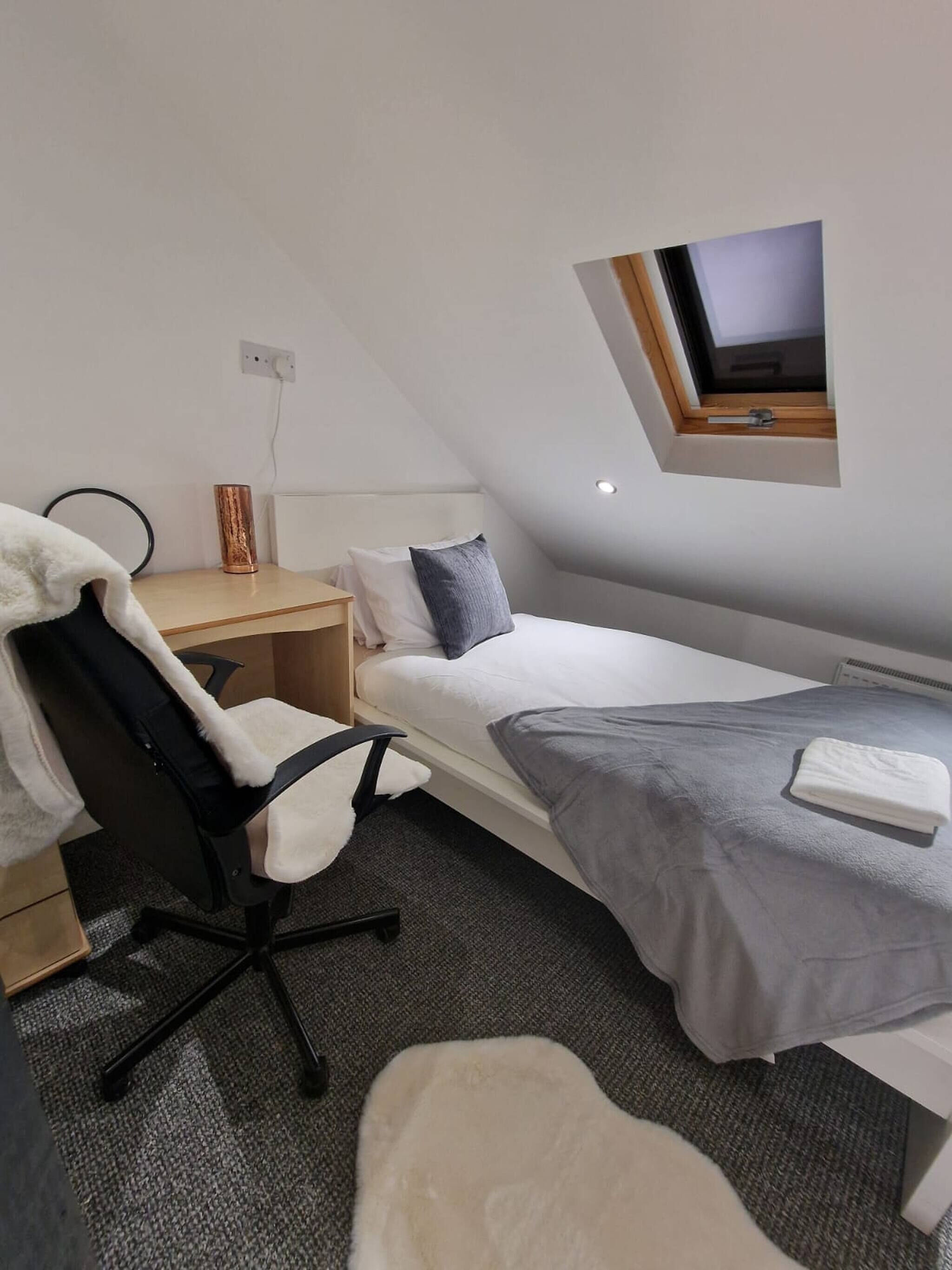 Une petite chambre moderne, équipée d’un lit simple, d’un bureau, d’une chaise, d’une petite table, d’une prise de courant et d’une fenêtre.