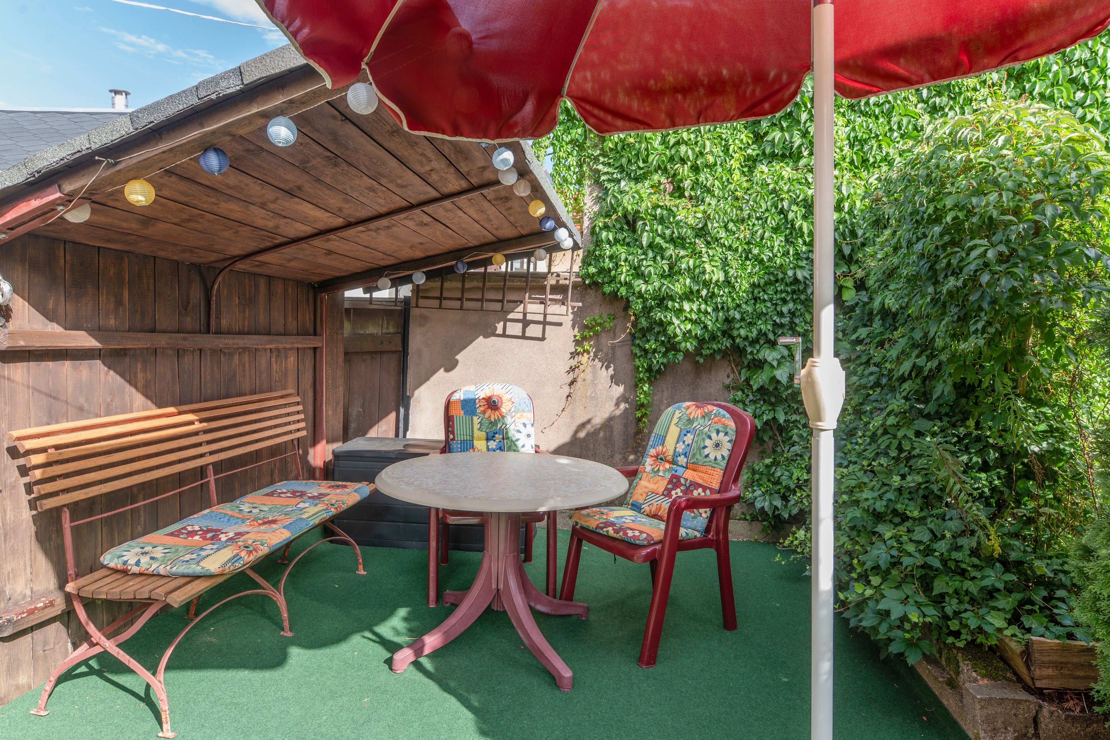 Een terras met een houten bankje, een tafel en stoelen, omringd door groen en een rode parasol.