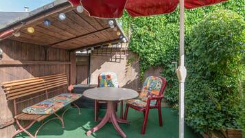 Een terras met een houten bankje, een tafel en stoelen, omringd door groen en een rode parasol.