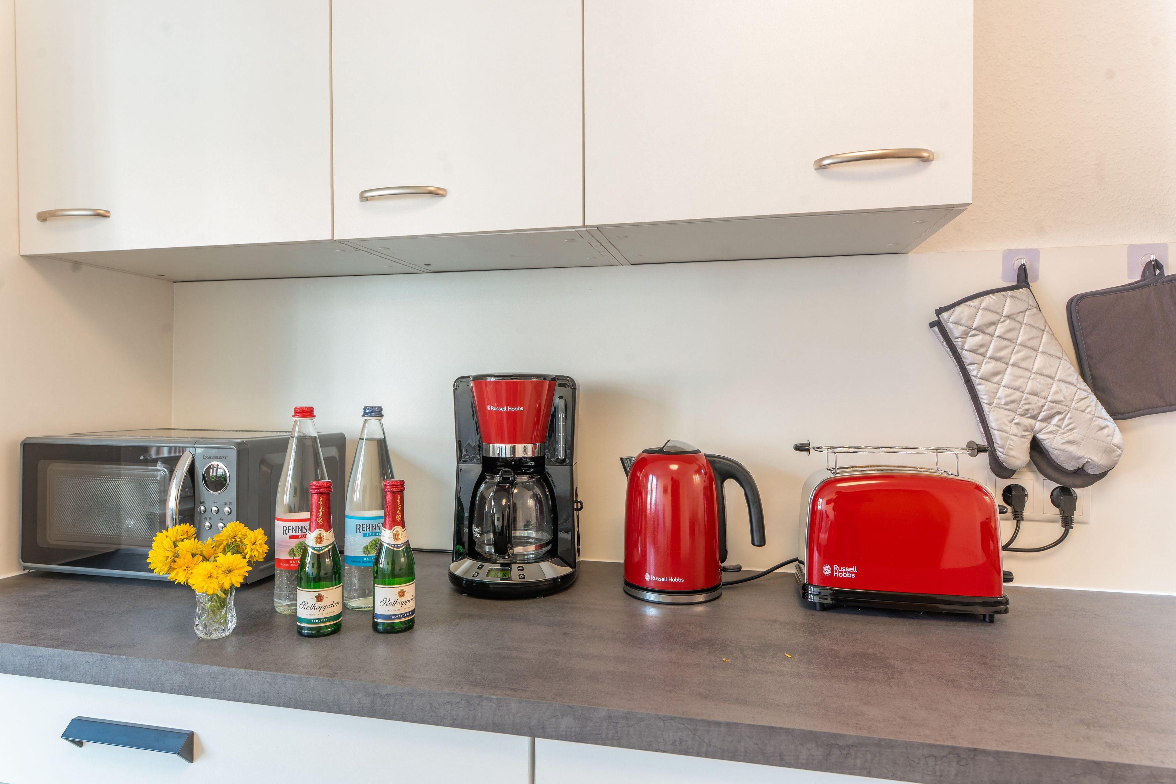 Un piano di lavoro in cucina con un forno a microonde, una macchina per il caffè, un bollitore e un tostapane. Si vedono anche due bottiglie di birra e un vaso con fiori.