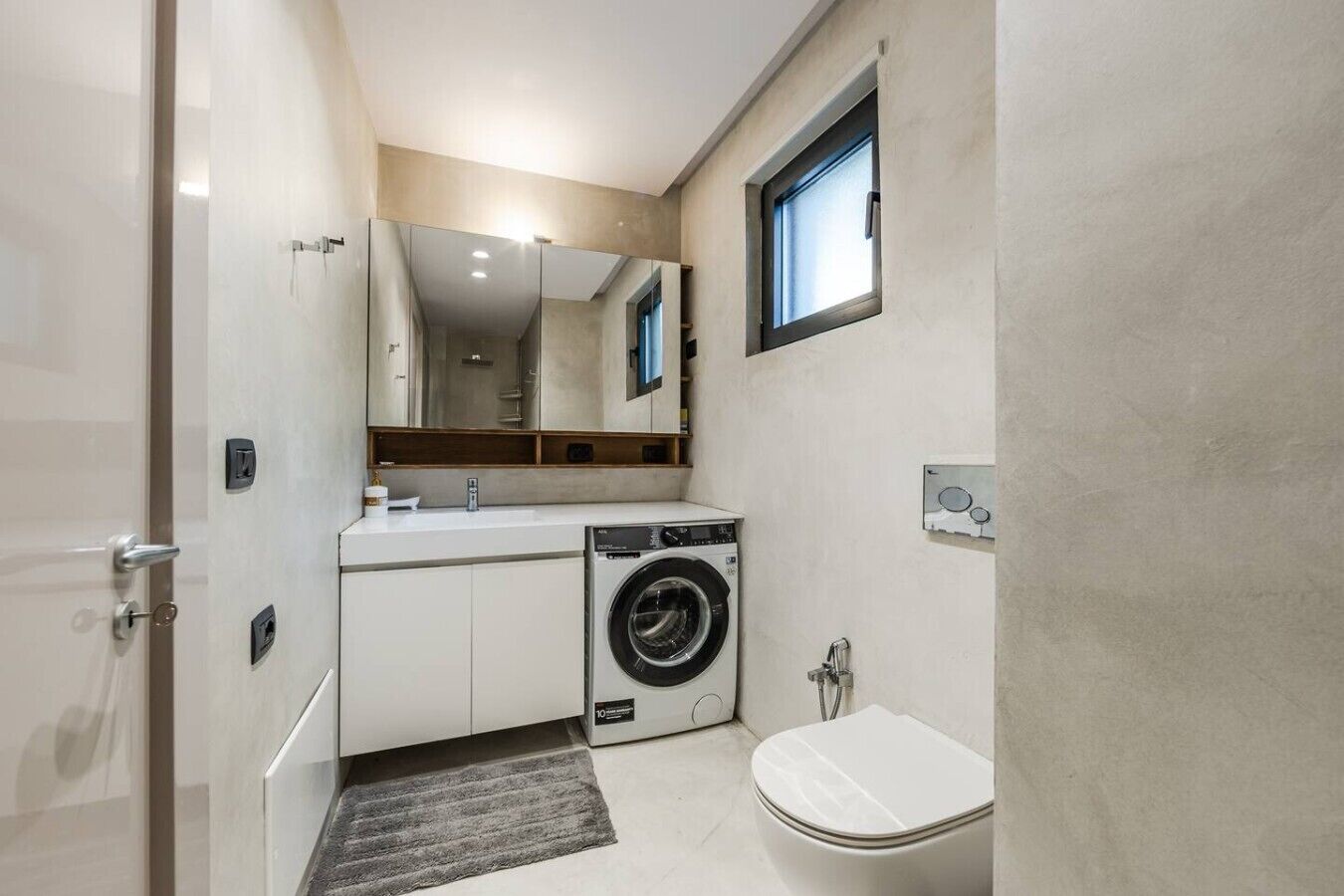 Une petite salle de bain équipée d’un lave-linge, de toilettes et d’un miroir au-dessus du lavabo.