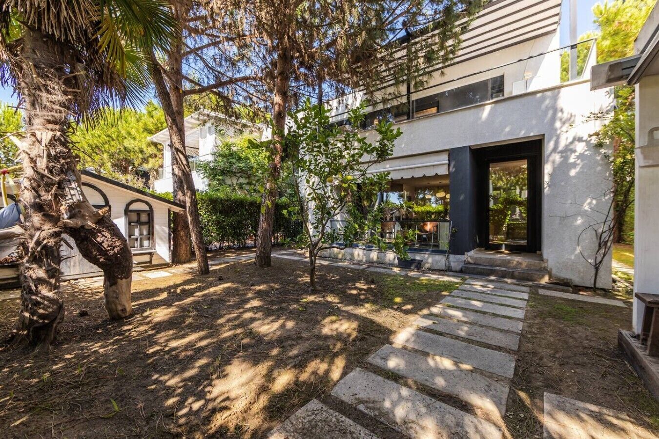 Un edificio moderno con un área de estar al aire libre techada, un sendero de piedra y varios árboles.