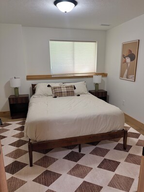 A bedroom with a bed, two bedside tables, a window with blinds, a wall-mounted light fixture, and a framed picture.