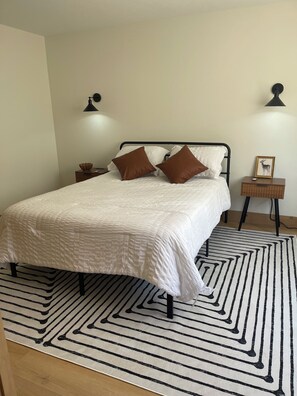 A neatly made bed with a white duvet and two brown pillows, a black and white striped rug, and two bedside tables with lamps and a framed picture.