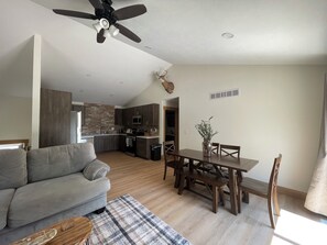 A modern living room with a sofa, dining table, and chairs, a kitchen in the background, and a ceiling fan.