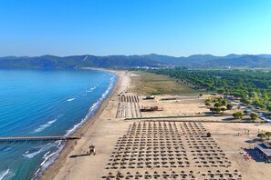 Vista aérea de una playa con hileras de sombrillas y sillas de playa, un muelle que se adentra en el mar, y colinas y edificios al fondo.