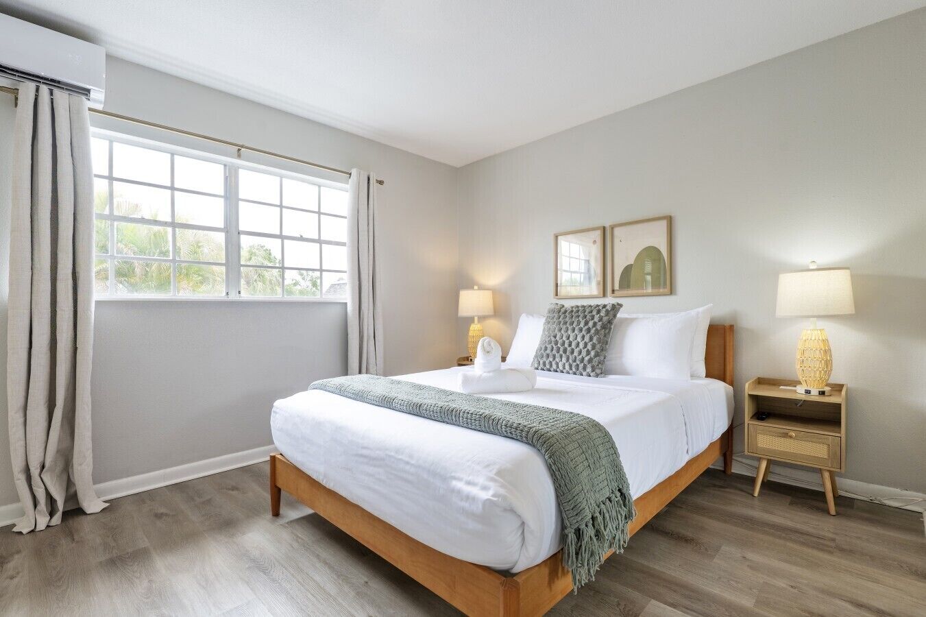 A hotel room with a large bed, bedside lamps, a window with curtains, and a wall-mounted air conditioner.