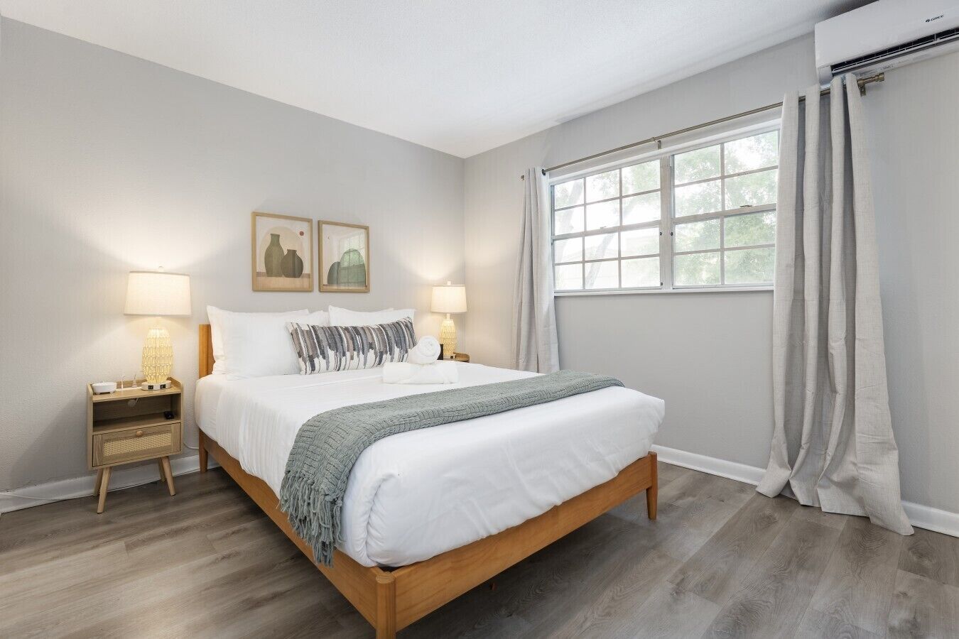 A bedroom with a wooden bed, two bedside tables with lamps, a window with curtains, and framed artwork on the wall.