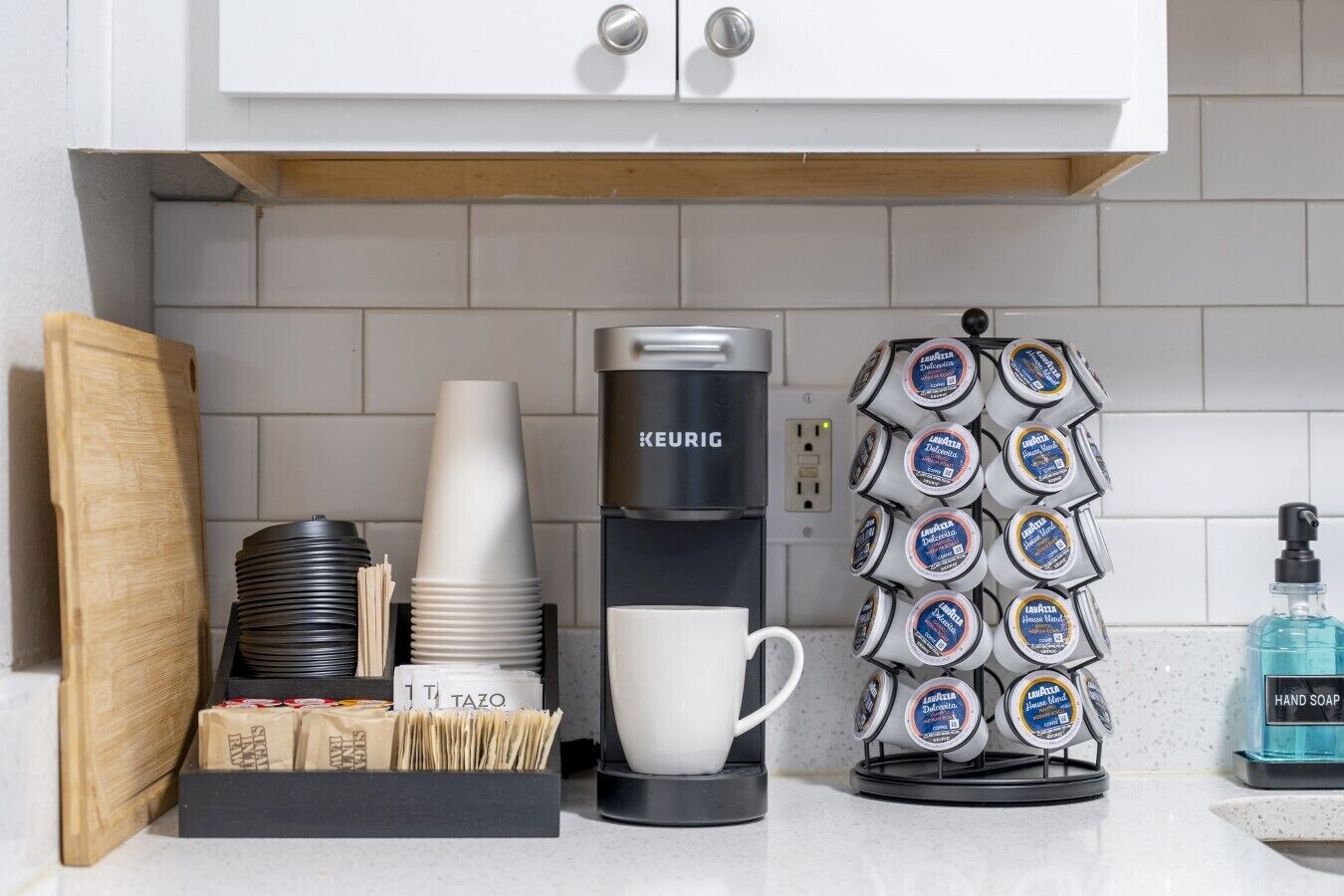 A coffee station with a Keurig coffee maker, disposable cups, a coffee mug, a paper cup holder, and a hand soap dispenser.