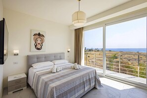 Une chambre avec un grand lit, une table de chevet, une commode, une lampe fixée au mur, un tableau et un balcon avec vue sur la mer.
