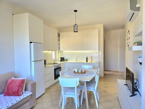 A compact kitchen with white cabinetry, a dining table with chairs, a sofa with a red pillow, and a TV on a stand. - Lura 2 Holiday Homes 181 (Gjiri Lalzit)