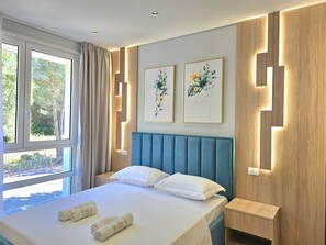 A modern hotel room with a large bed, two wall-mounted artworks, a window with curtains, and a bedside table. - White Shell Apartments (Gjiri Lalzit)
