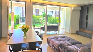 Een moderne woonkamer met een eethoek, een bank en uitzicht op een tuin door grote glazen deuren.