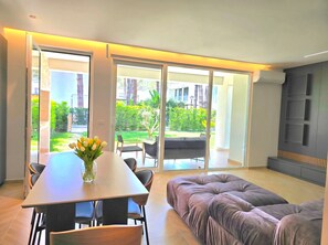 A modern living room with a dining area, a sofa, and a view of a garden through large glass doors. - Relax Apartment By The Sea (Gjiri Lalzit)
