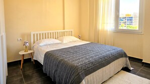 Quarto com cama, mesa de cabeceira com abajur, janela com cortinas e vista para prédios.