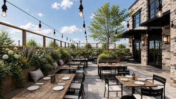 An outdoor dining area with wooden tables and chairs, string lights, and a stone building in the background.