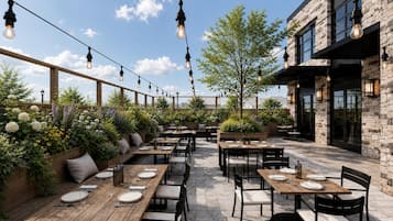 An outdoor dining area with wooden tables and chairs, string lights, and a stone building in the background.