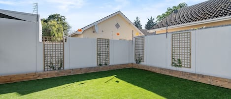 Une cour arrière clôturée avec une pelouse artificielle, une clôture en bois et un bâtiment doté d’un toit en tuiles.