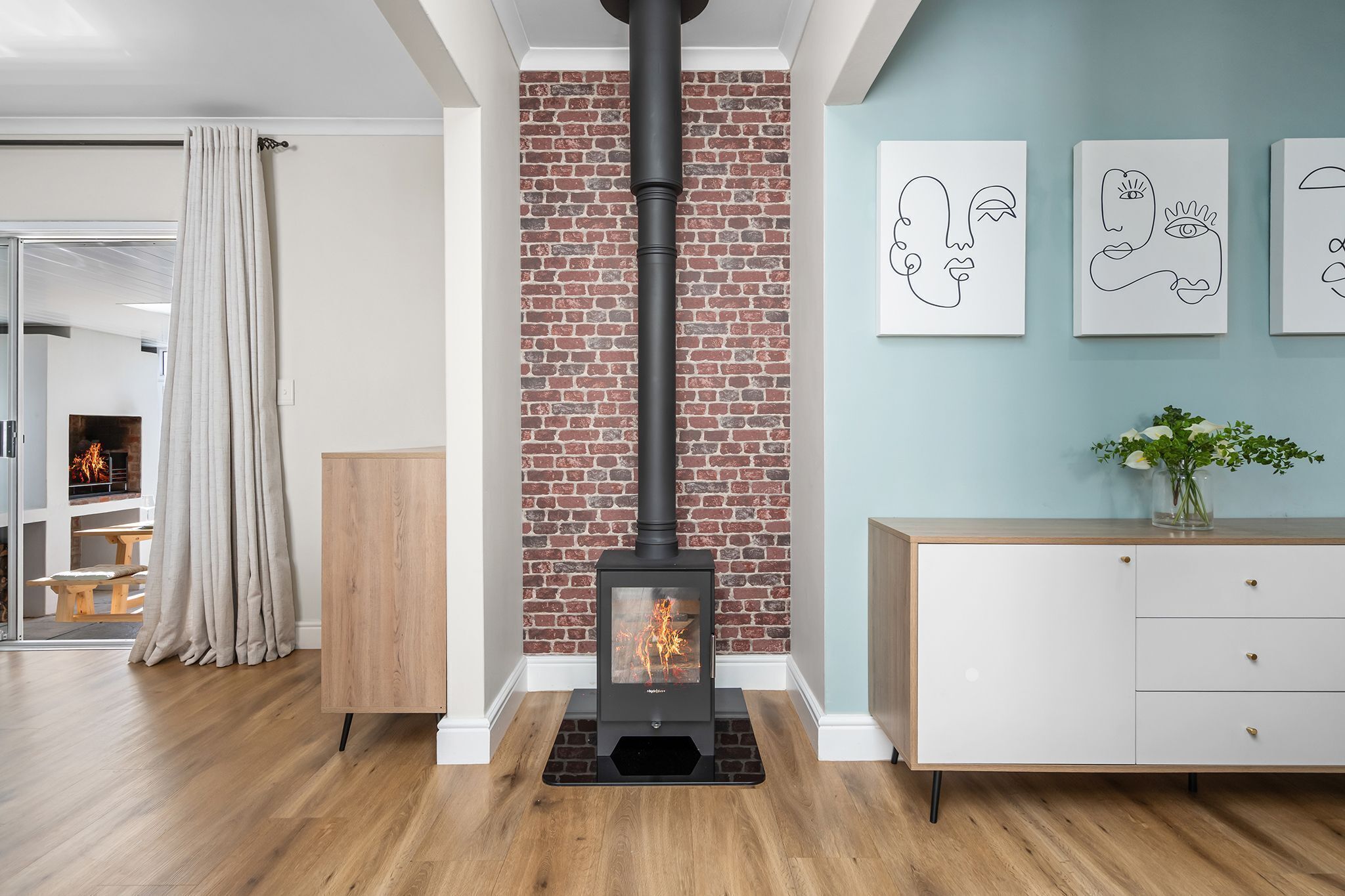 A modern living room with a brick wall, a black wood-burning stove, a white cabinet, and abstract art on the wall.