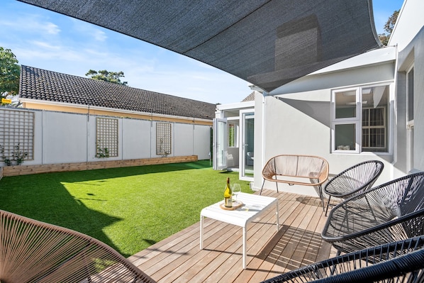 A patio with outdoor seating, a table with a bottle and glass, and a view of a house with a white exterior and a tiled roof.